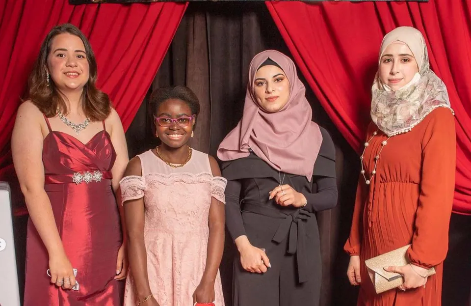 Quattro studentesse con vestiti colorati e hijab si trovano davanti a una tenda rossa nei pressi della John Barsby Secondary School.