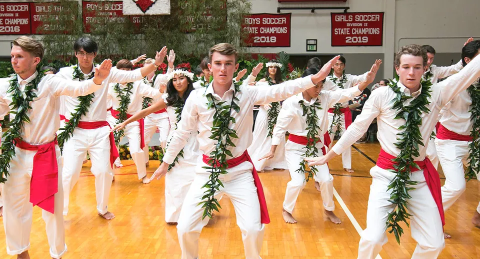 Studenti della Hawaii Preparatory Academy eseguono una danza tradizionale con costumi bianchi e rossi su un palco.