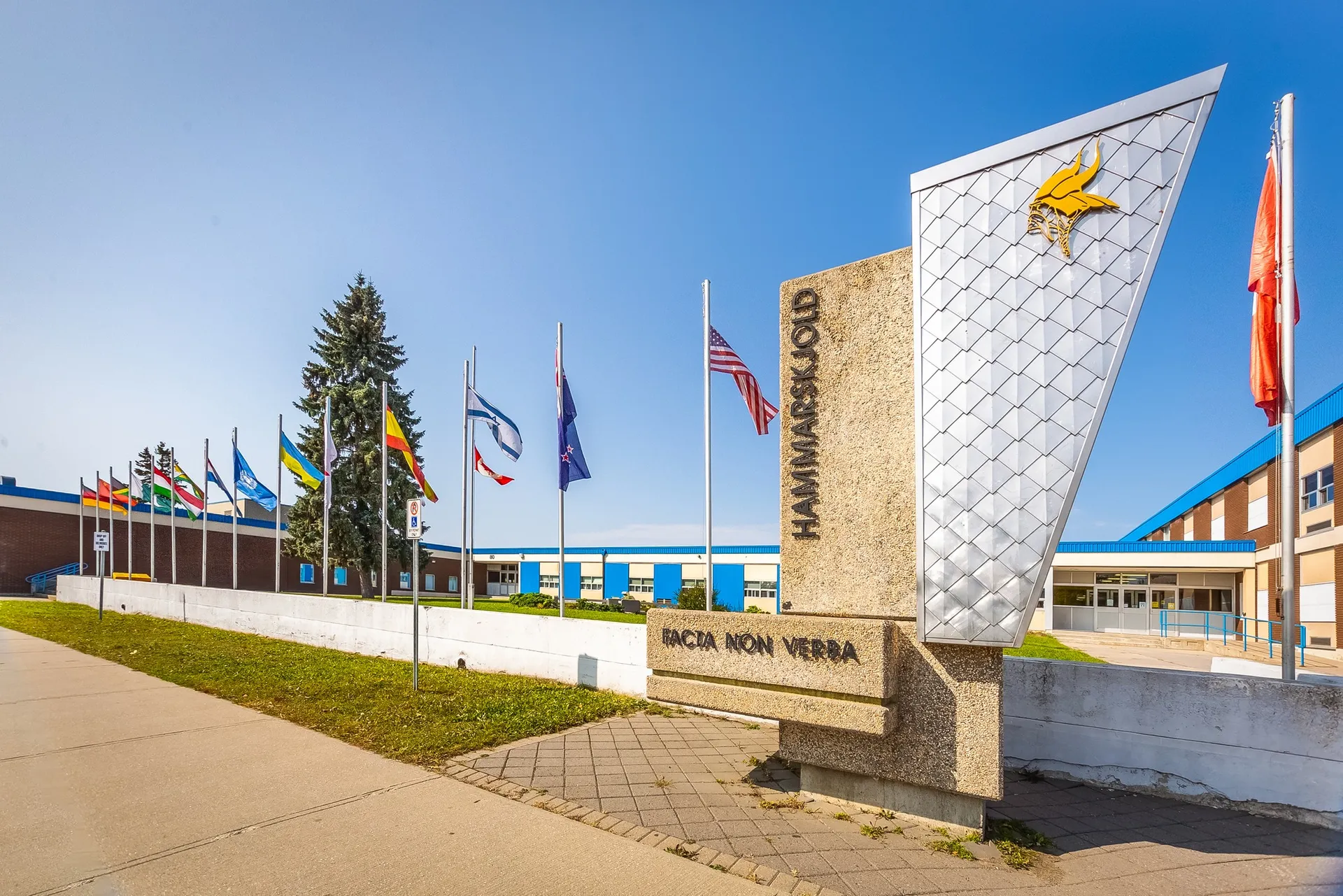 L'edificio scolastico moderno a più piani della Hammarskjold High School, con facciata decorativa, è circondato da bandiere e da un viale pavimentato.