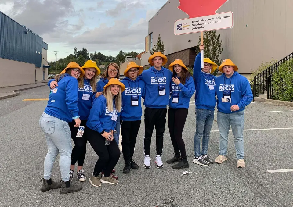 Un gruppo di studenti della Exploits Valley High School si trova davanti a un cantiere con camicie blu ed elmetti di protezione.