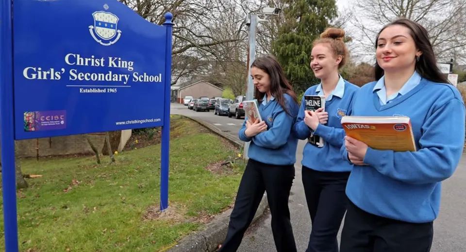 Tre studentesse sono davanti a un cartello della Christ King Secondary School con alberi e un prato sullo sfondo.