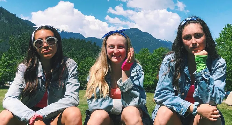 Tre studentesse della Pemberton Secondary School siedono insieme all'aperto con montagne e bosco sullo sfondo.
