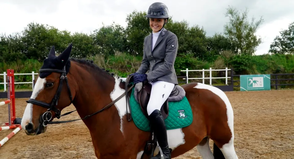 Una studentessa della Rockport School cavalca un cavallo marrone in un maneggio all'aperto con alberi e una recinzione.