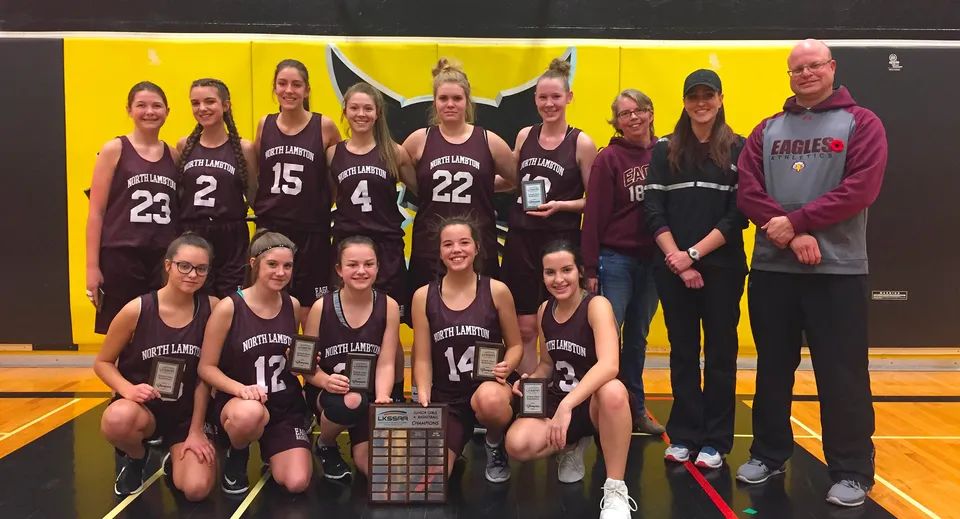 Un gruppo di giovani giocatrici di basket posa con un trofeo davanti a uno sfondo giallo nel campus della North Lambton Secondary School.