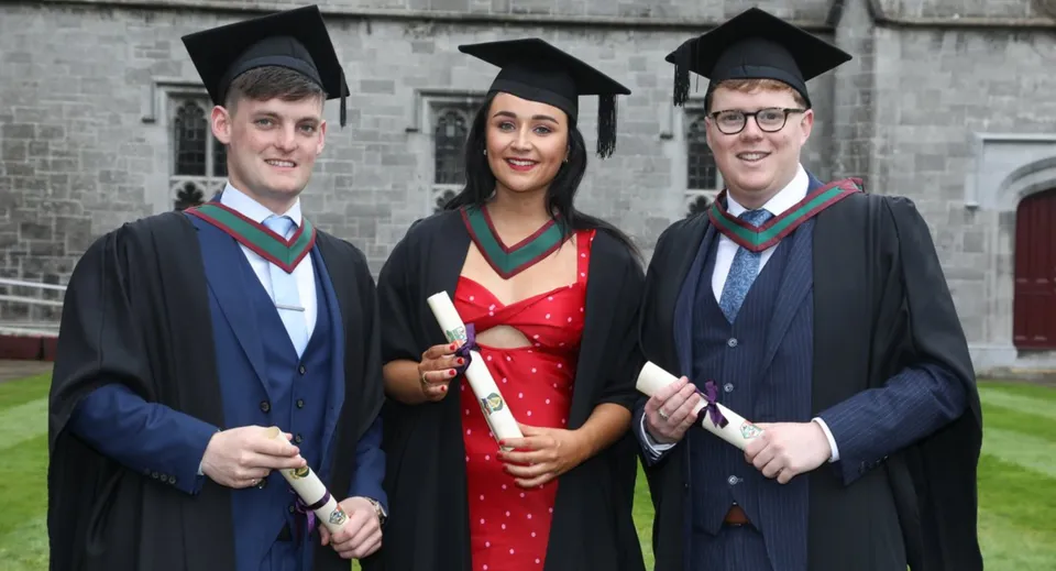 Tre studenti del Holy Rosary College in toga da diploma davanti a un edificio in mattoni con i diplomi in mano.