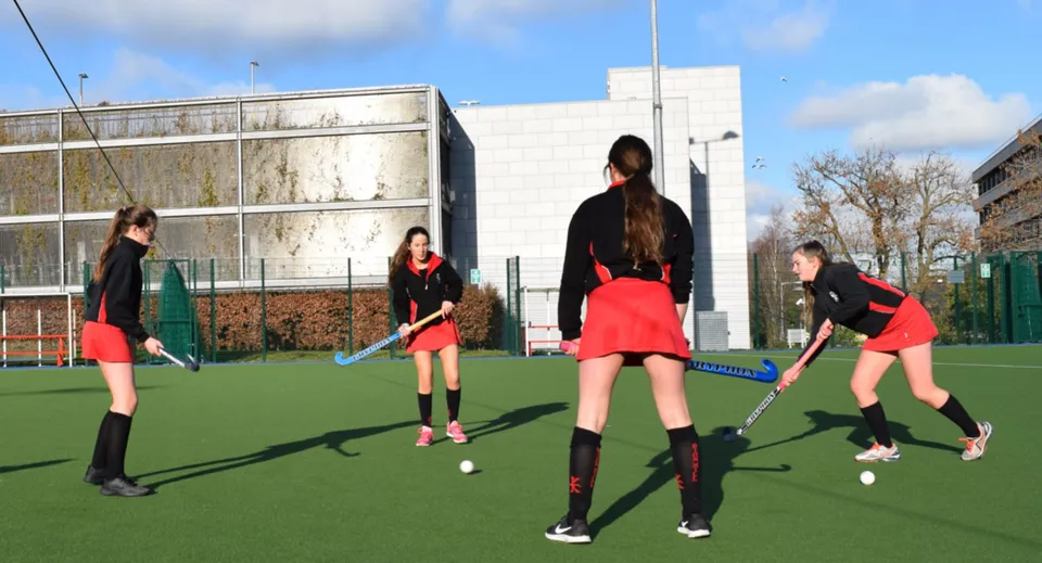 Studentesse del Dominican College Sion Hill giocano a hockey su prato sul campo in erba sintetica con un edificio scolastico sullo sfondo.