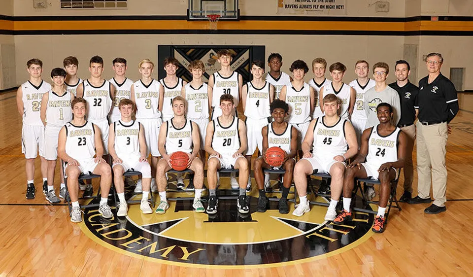 La squadra di basket della Maur Hill-Mount Academy posa insieme agli allenatori sul campo da gioco davanti al logo della squadra.