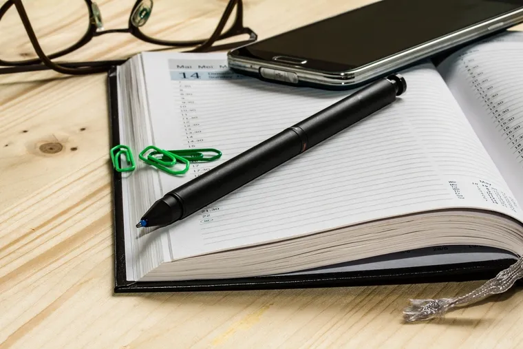 Aufgeschlagener Terminkalender mit Kugelschreiber, Smartphone, Brille und grünen Büroklammern
