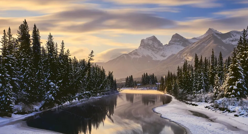 Un paesaggio invernale innevato in Canada con un fiume, pini e montagne innevate sullo sfondo.