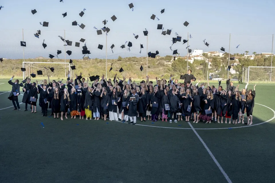 Un grande gruppo di diplomati e diplomate della Laude Lady Elizabeth School lancia in aria i tocchi da diploma sul campo sportivo.