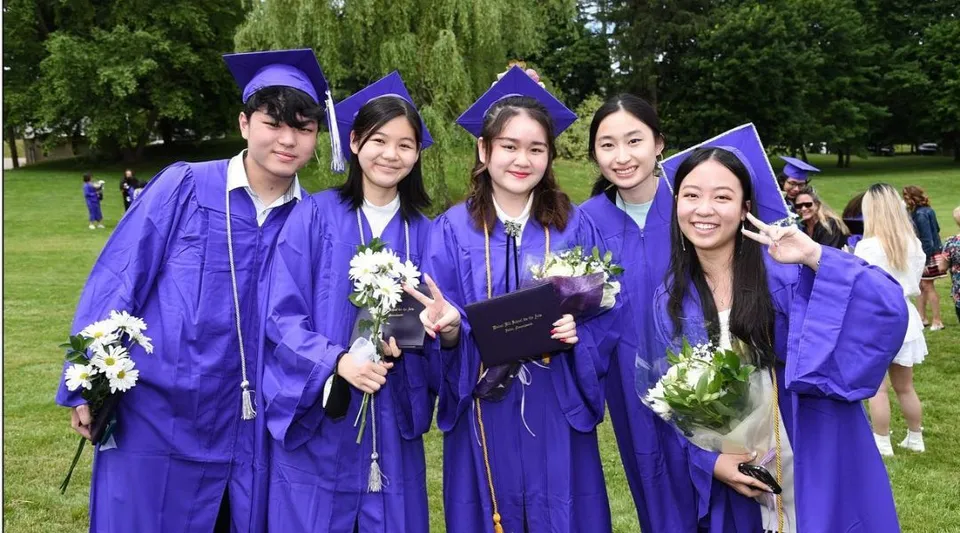 Un gruppo di giovani donne della Walnut Hill School for the Arts si trova su un prato verde con fiori, indossando toghe viola per la cerimonia di diploma.