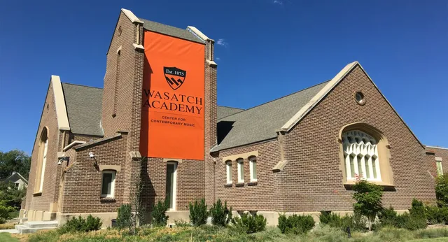 Das große Backsteingebäude mit einem orangefarbenen Banner der Wasatch Academy vor klarem blauem Himmel und grüner Umgebung.