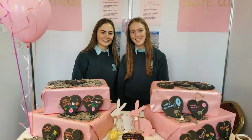 Due studentesse della St. Louis Secondary School stanno davanti a uno sfondo rosa con due grandi torte decorate con cuori in primo piano.