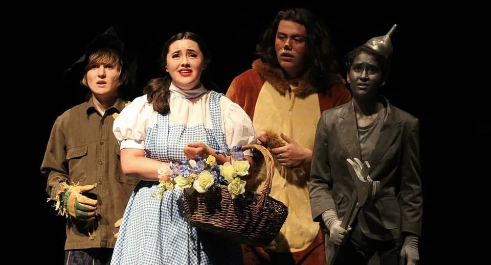 Un gruppo di studenti della Esquimalt High School posa in costumi storici con fiori e cesti davanti a uno sfondo scuro.