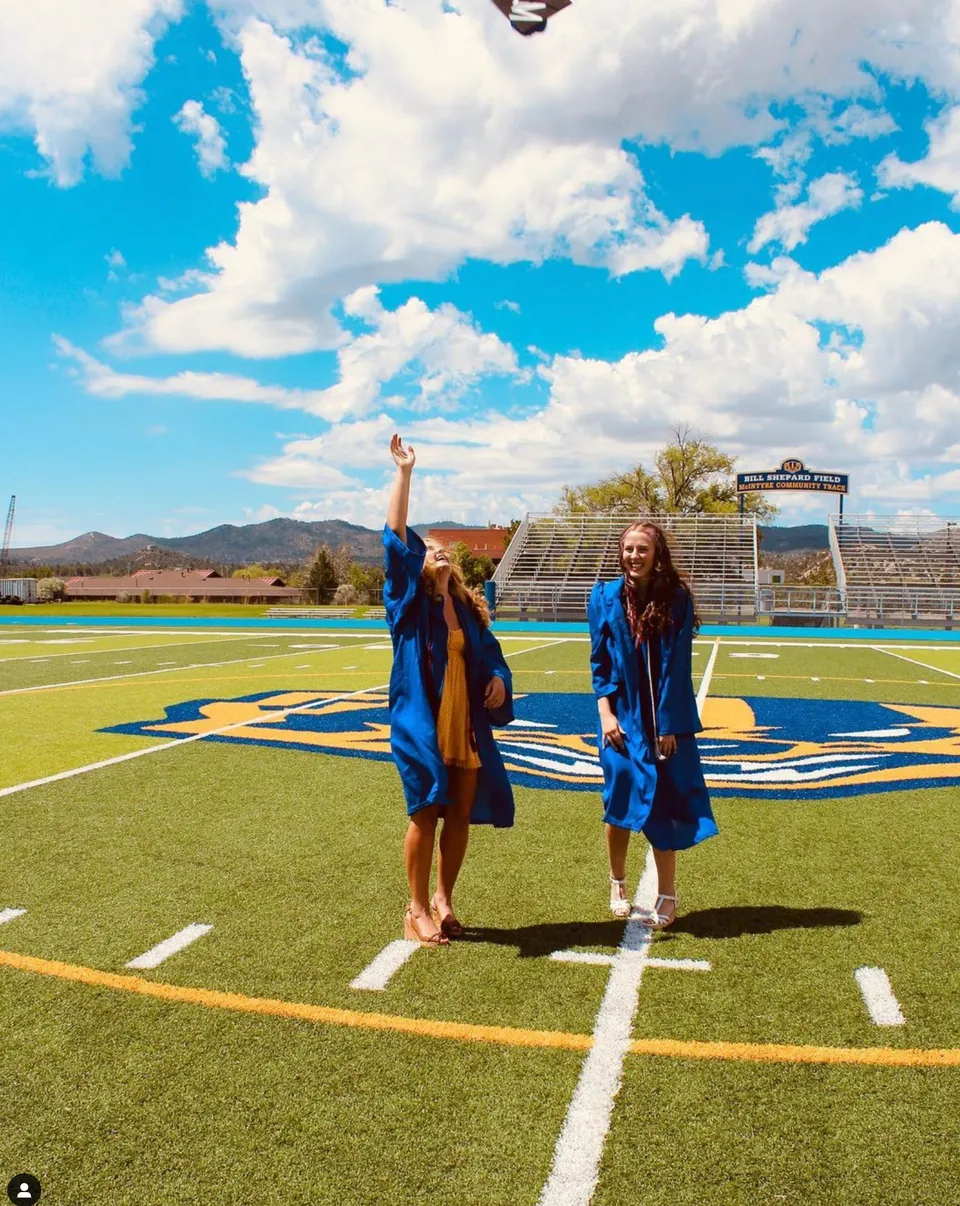 Due diplomati con toghe blu sono in piedi sul campo sportivo della Prescott High School davanti a montagne e un cielo nuvoloso.