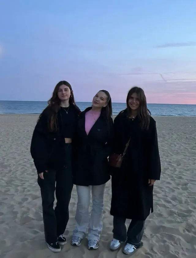 Tre amiche in cappotto scuro posano su una spiaggia inglese al tramonto
