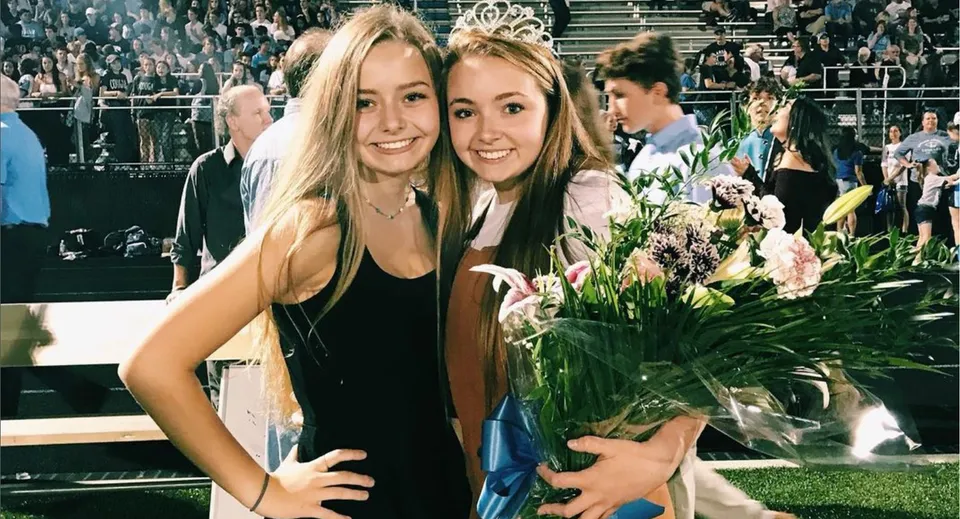 Due studentesse della Lansing Catholic High School tengono insieme un mazzo di fiori e posano con la folla su un palco sullo sfondo.