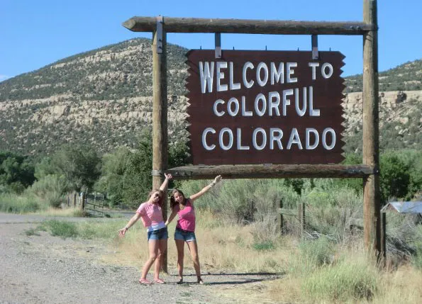 Dos chicas sonrientes posan bajo un cartel de madera con el texto Welcome to Colorful Colorado