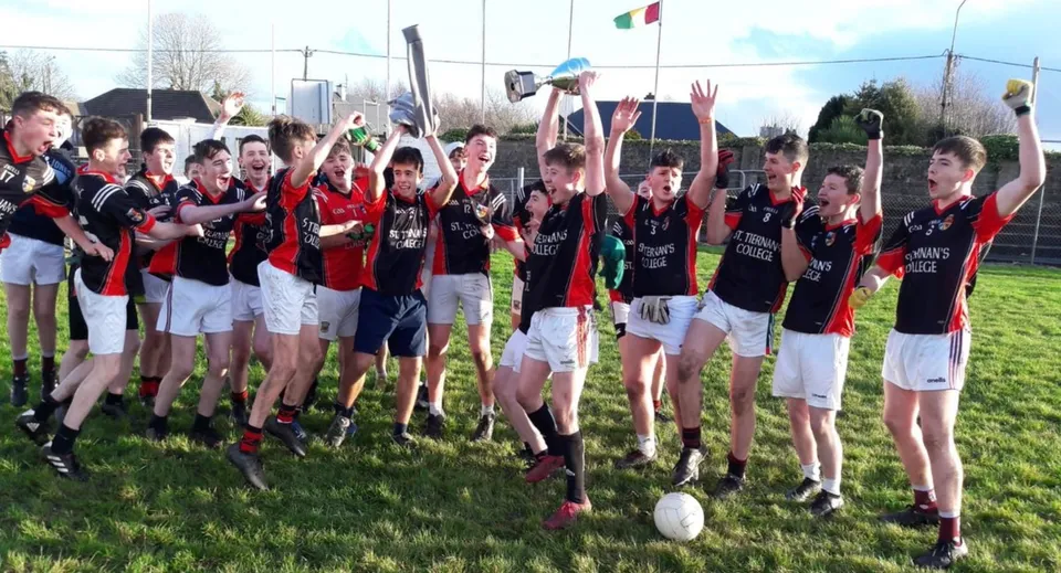 Studenti del St. Tiernan’s College festeggiano insieme nelle loro divise sportive su un campo sportivo con uno stadio sullo sfondo.