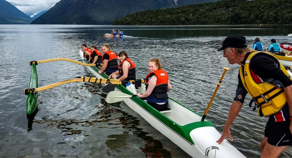 Studenti della Riccarton High School seduti con giubbotti di salvataggio su una barca in un lago tra montagne e foreste.