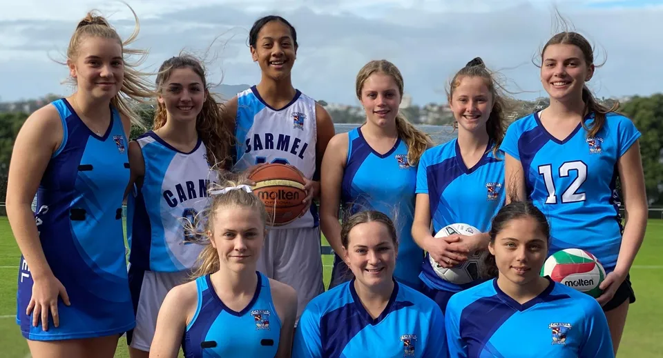 Un gruppo di giovani giocatrici di basket in maglia blu posa su un campo sportivo del Carmel College.