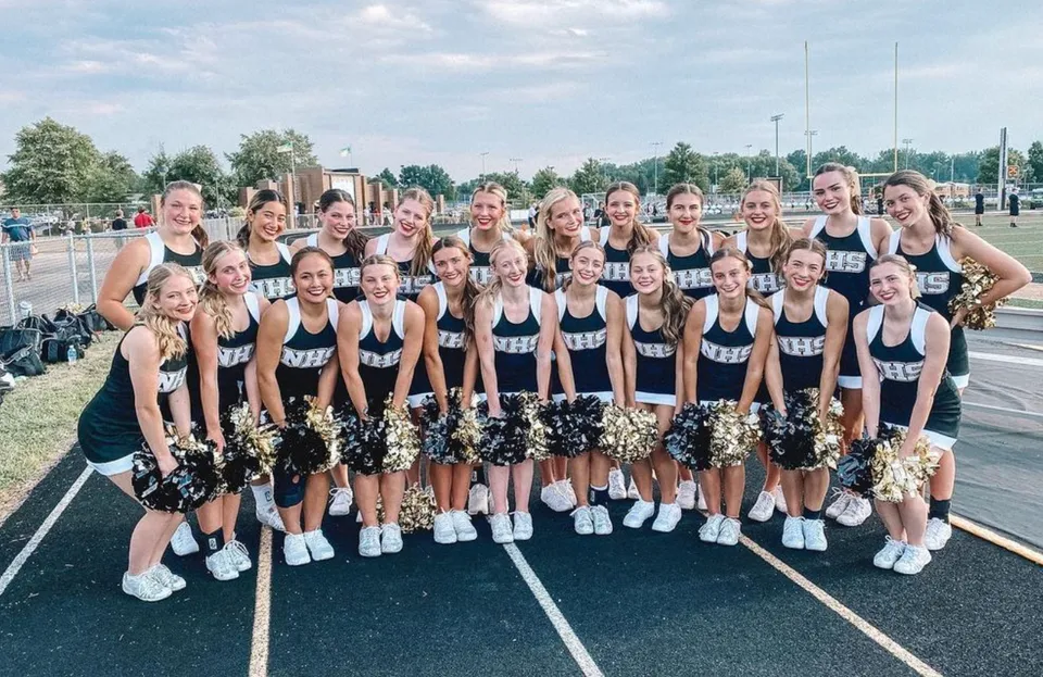 Un gruppo di cheerleader della Mt. Vernon High School posa insieme sul campo sportivo con edifici e alberi sullo sfondo.