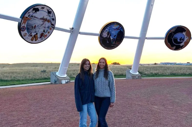 Dos jóvenes sonríen al aire libre al atardecer bajo una estructura metálica blanca