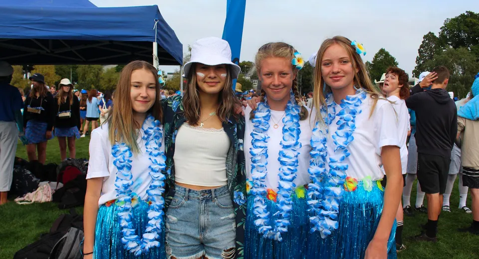 Un gruppo di studentesse della Rangiora High School indossa collane di fiori colorate davanti a una tenda blu durante un evento all'aperto.