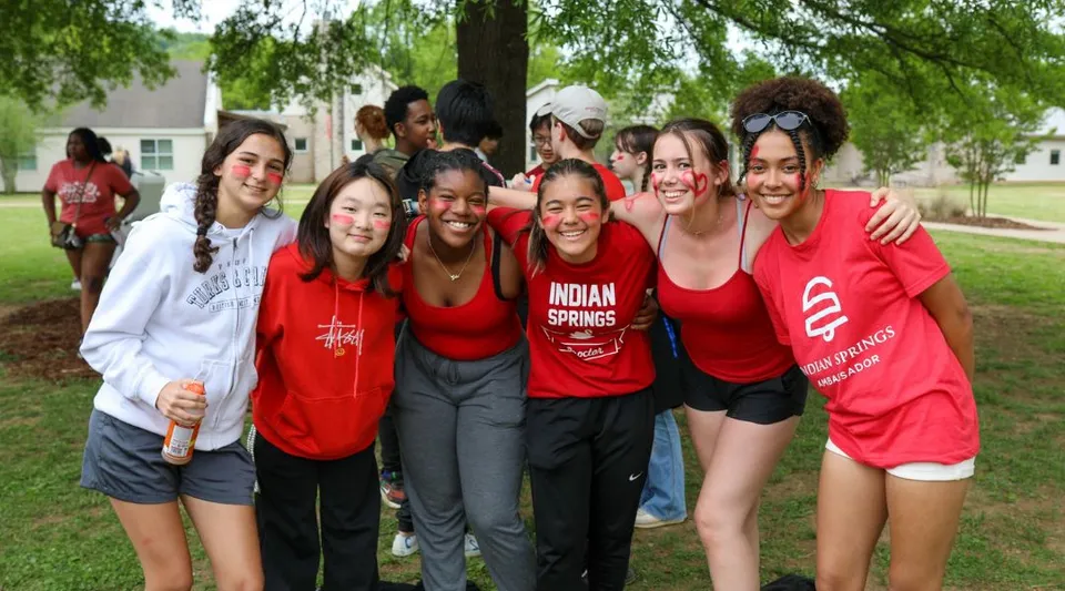 Un gruppo di giovani donne con le magliette rosse della Indian Springs School si trova all'aperto su un prato con alberi sullo sfondo.