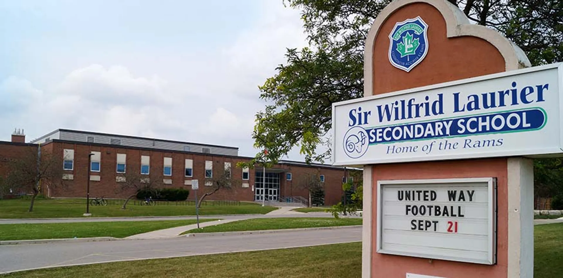 Il cartello per un evento di football di United Way davanti all'edificio in mattoni della Sir Wilfred Laurier Secondary School è circondato da alberi e prato.