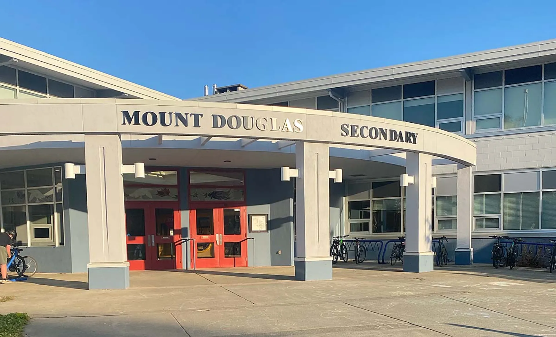L'edificio scolastico moderno a due piani della Mount Douglas Secondary School con porte d'ingresso rosse e cielo blu sullo sfondo.