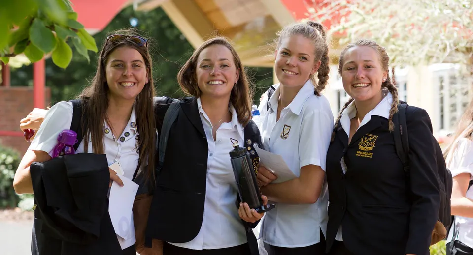 Quattro studentesse in uniforme sono in piedi insieme davanti a un edificio in mattoni nel campus della Whangarei Girls High School.
