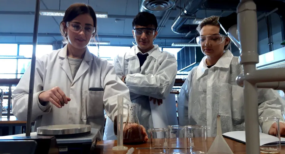Tre persone in camice da laboratorio bianco stanno intorno ad apparecchiature scientifiche nel laboratorio della Ballinteer Community School.