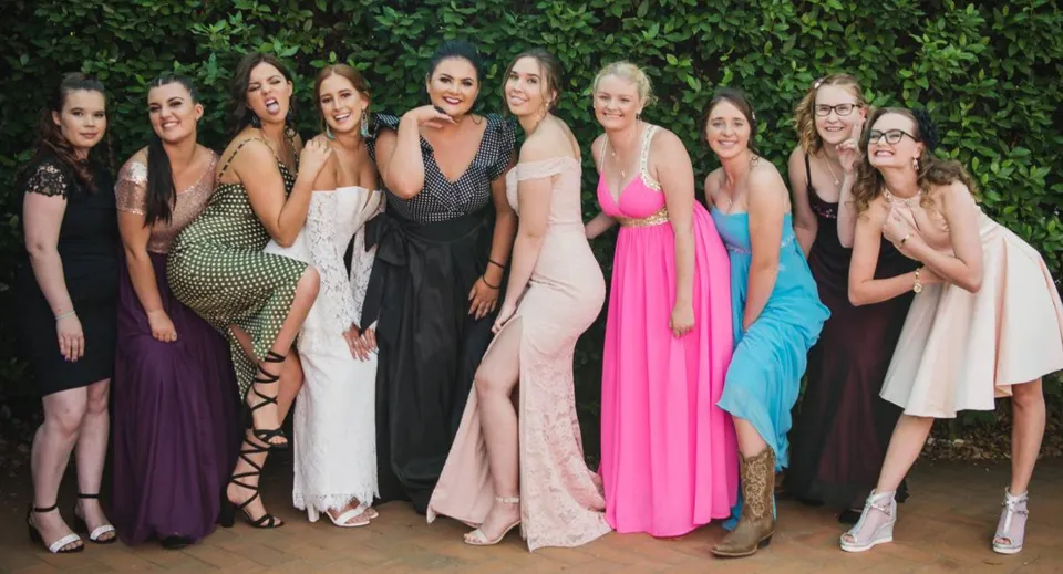 Un gruppo di studentesse della Maleny State High School posa davanti a un fogliame verde in vari abiti eleganti.