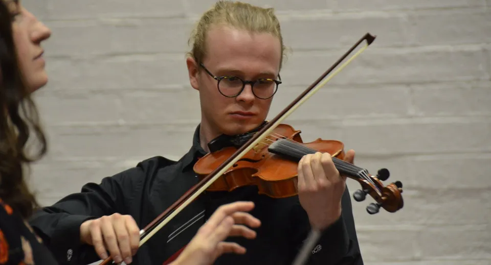 Uno studente di The High School suona concentrato il violino davanti a una parete di mattoni bianchi.