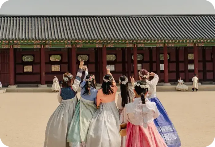 Ragazze in hanbok, di spalle, fanno il segno della pace davanti a un palazzo coreano