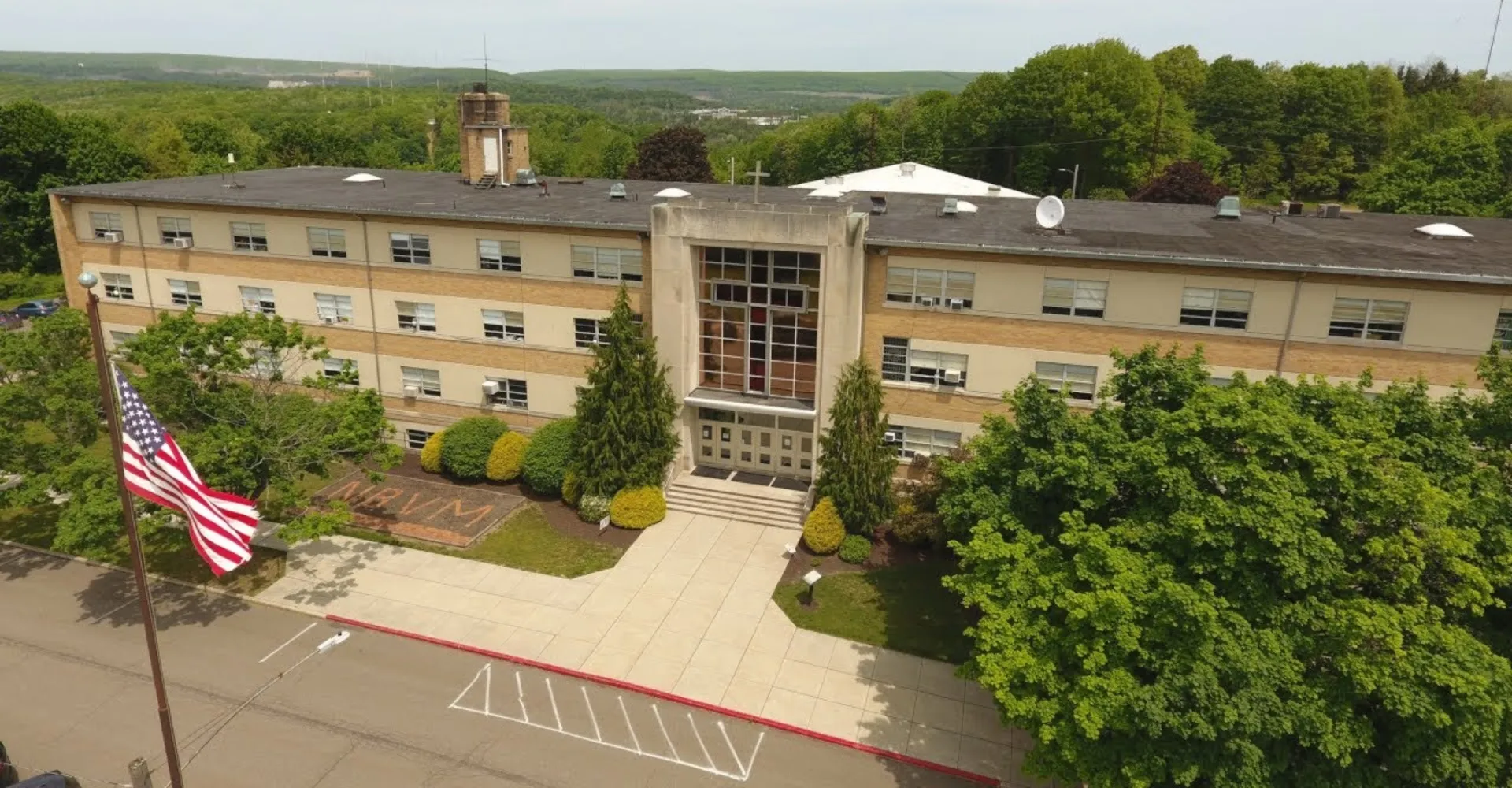 L'edificio principale a più piani della Nativity BVM High School, dotato di una torre e bandiere americane, è circondato da alberi.