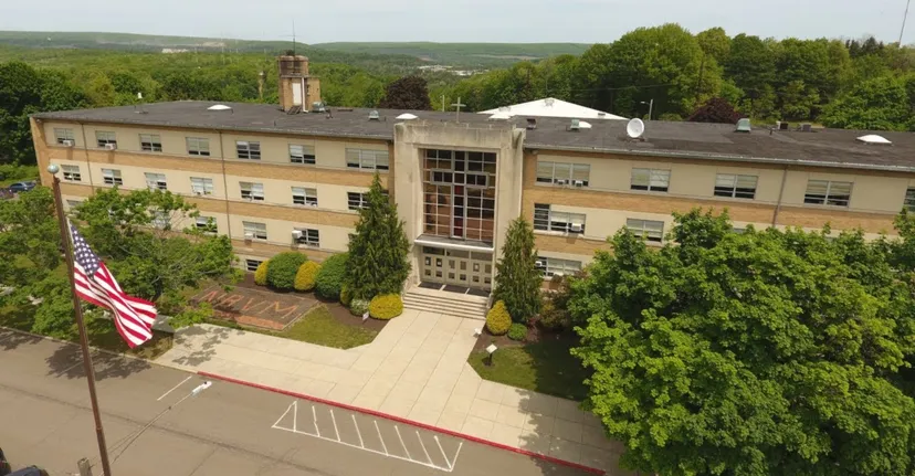 L'edificio principale a più piani della Nativity BVM High School, dotato di una torre e bandiere americane, è circondato da alberi.