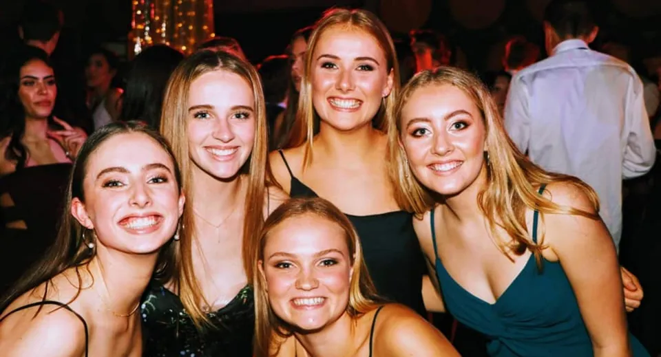 Studentesse della Westlake Girls High School posano insieme a una festa con luci colorate sullo sfondo.