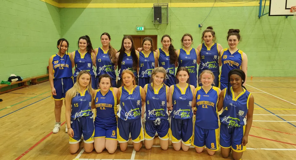 Un gruppo di giovani giocatrici di basket in maglia blu e gialla posa nella palestra della Mountmellick Community School.