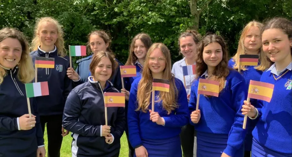 Studentesse della Ursuline Secondary School (Cork) si trovano all'aperto in uniforme blu e tengono in mano diverse bandiere nazionali.