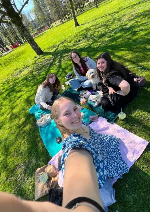 Lorena macht ein Selfie mit Freundinnen und einem kleinen Hund beim Picknick in einem sonnigen Park in Irland