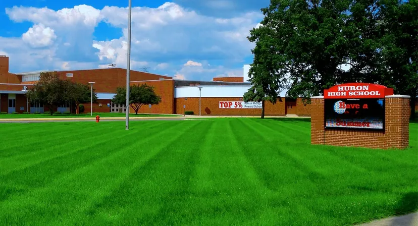 L'edificio della Huron High School si trova dietro un prato verde ben curato sotto un cielo blu con nuvole bianche.