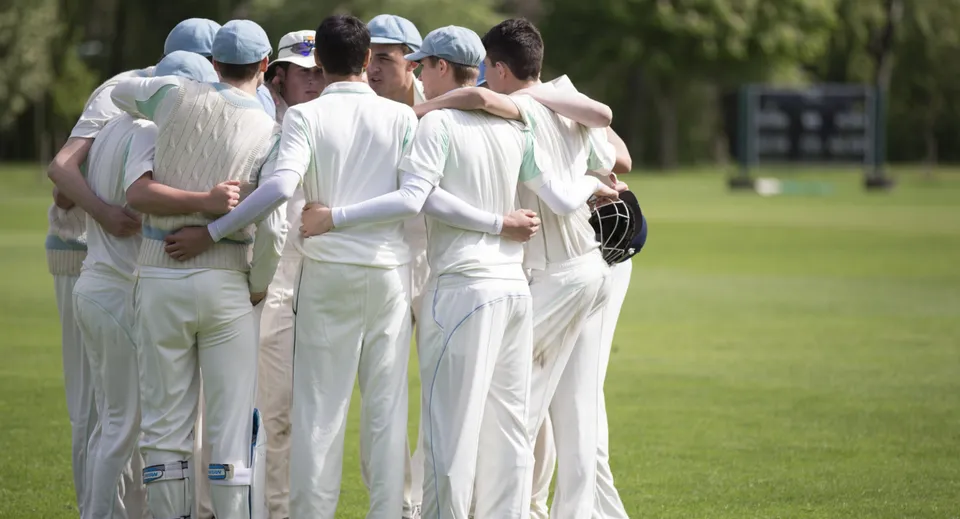 Un gruppo di studenti della Harrow School in divisa bianca da cricket si trova su un campo d'erba davanti a un tabellone segnapunti.