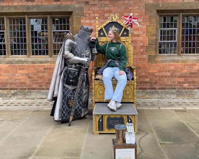 Chica sentada en un trono dorado junto a una estatua viviente vestida de caballero medieval
