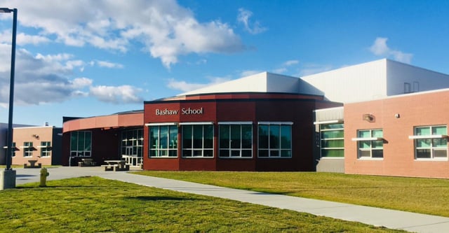 L'edificio scolastico moderno a più piani della Bashaw School con un ingresso rosso si trova su un prato sotto un cielo blu con nuvole bianche.