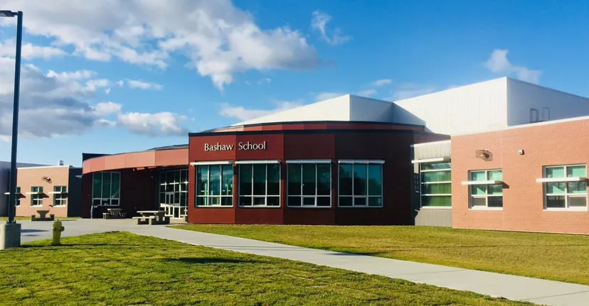 L'edificio scolastico moderno a più piani della Bashaw School con un ingresso rosso si trova su un prato sotto un cielo blu con nuvole bianche.