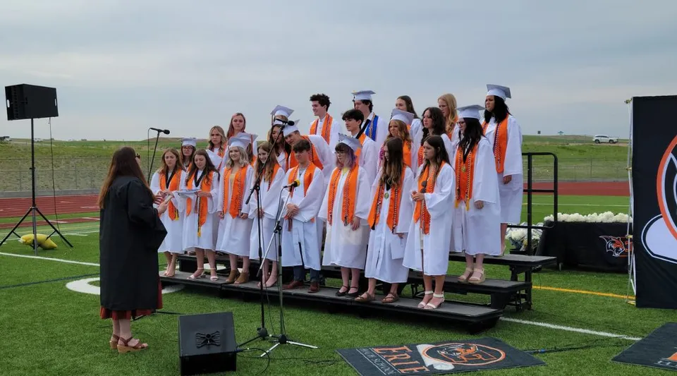Un gruppo di studentesse della Erie High School sta in piedi in toghe bianche e arancioni su un palco all'aperto davanti ad altoparlanti.