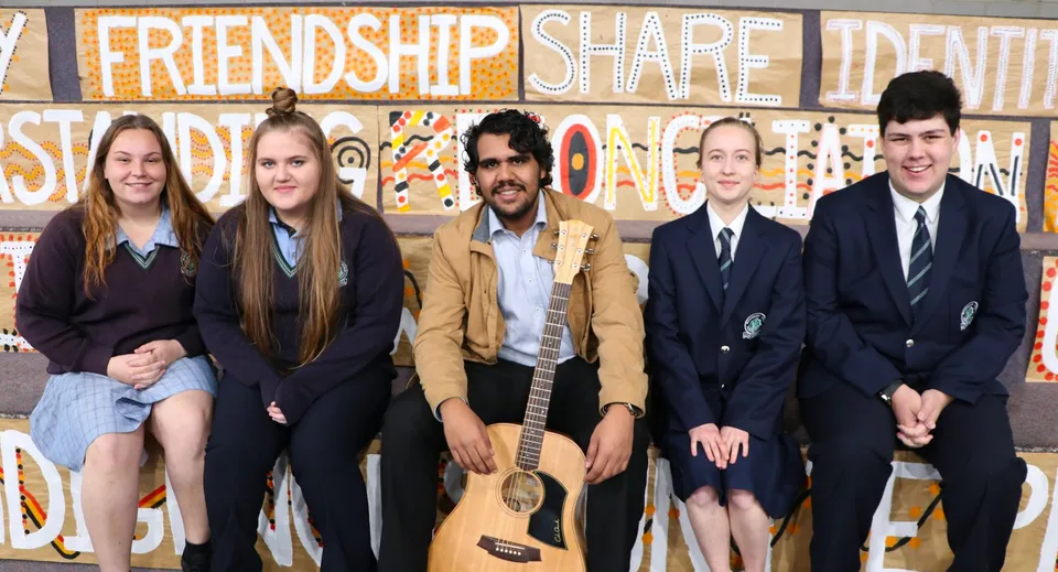 Un gruppo di studenti e studentesse si trova davanti a un murales colorato con le parole Friendship, Share e Identity nel campus della Modbury High School.