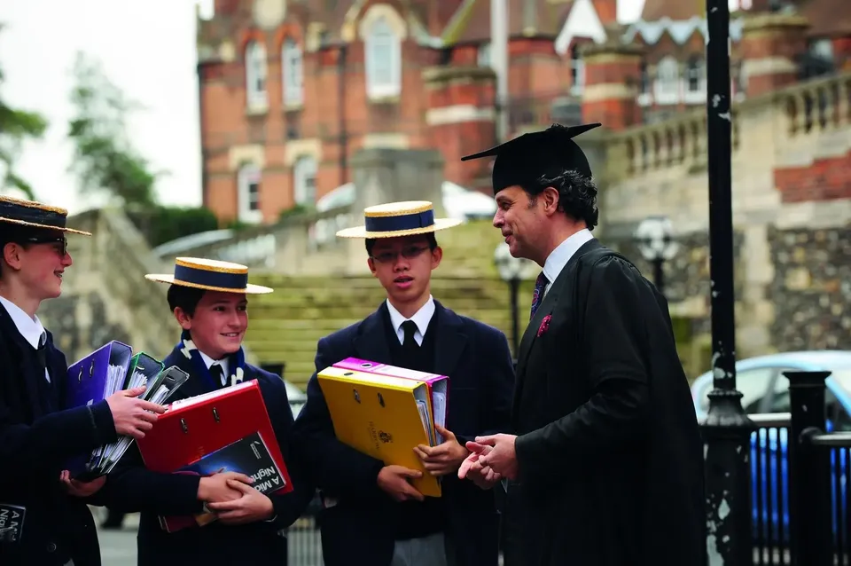 Un gruppo di studenti della Harrow School in abito da cerimonia di diploma si trova su una strada davanti a un edificio in mattoni tenendo in mano libri e documenti.